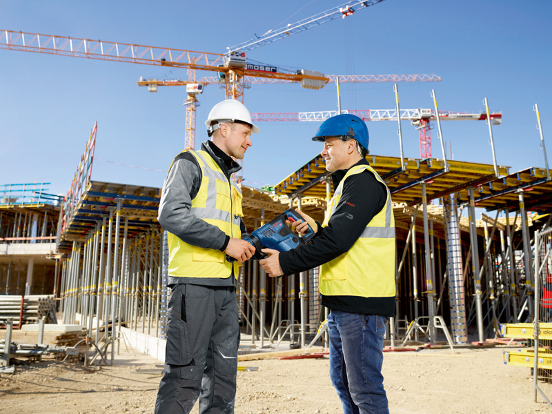 Zwei Personen in Sicherheitsausrüstung besprechen auf einer Baustelle ein Elektrowerkzeug. Anwendungsbild