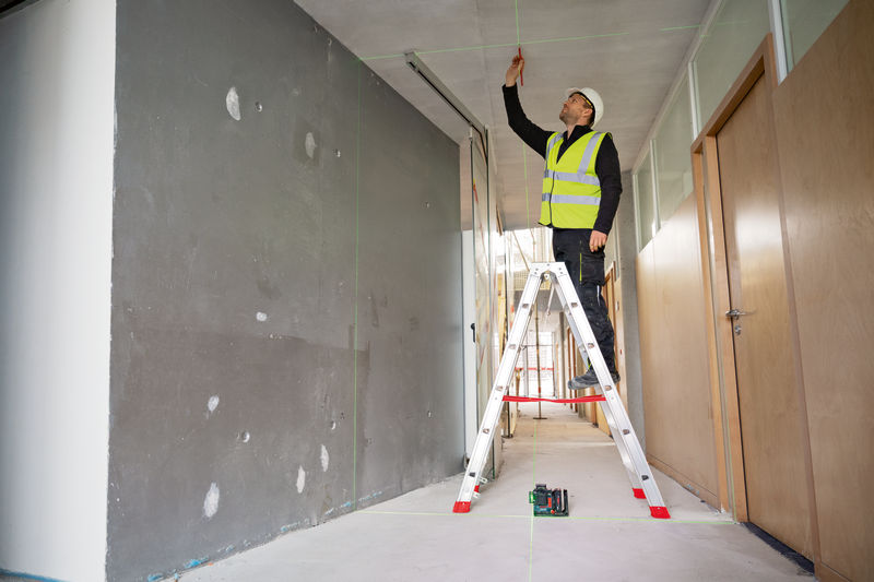 Eine Person mit Sicherheitsausrüstung markiert eine Decke mit einem Lasernivelliergerät. Anwendungsbild