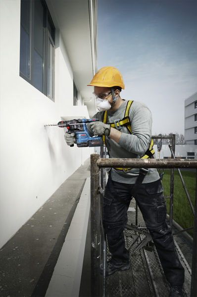 Ein Arbeiter mit Sicherheitsausrüstung bohrt auf einem Gerüst stehend in eine Gebäudefassade. Anwendungsbild