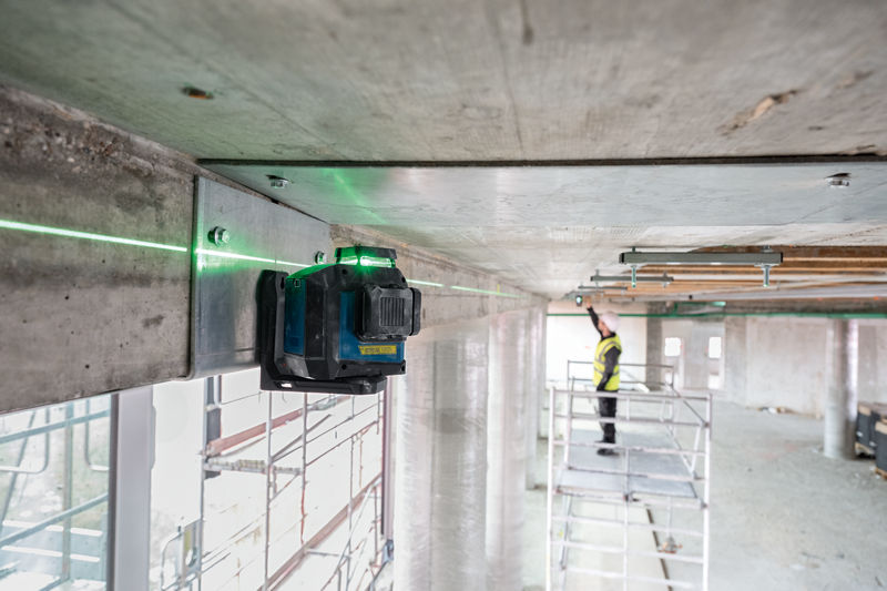 Ein Lasernivelliergerät projiziert eine grüne Linie über die Decke, eine Person mit Sicherheitsausrüstung passt die Einstellung an. Anwendungsbild