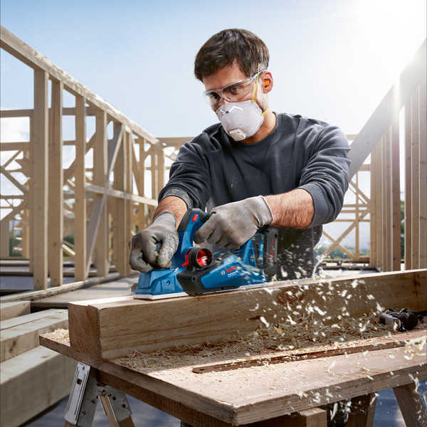 Eine Person mit Sicherheitsausrüstung hobelt auf einer Baustelle einen Holzbalken. Anwendungsbild