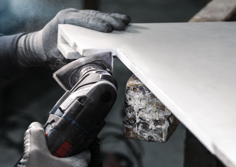 Eine Person mit Sicherheitsausrüstung schleift mit einem Multifunktionswerkzeug die Kante einer Arbeitsplatte aus Massivmaterial. Anwendungsbild