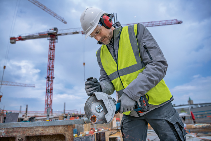 Eine Person mit Schutzausrüstung schneidet auf einer Baustelle Metall mit einem Akku-Winkelschleifer. Anwendungsbild
