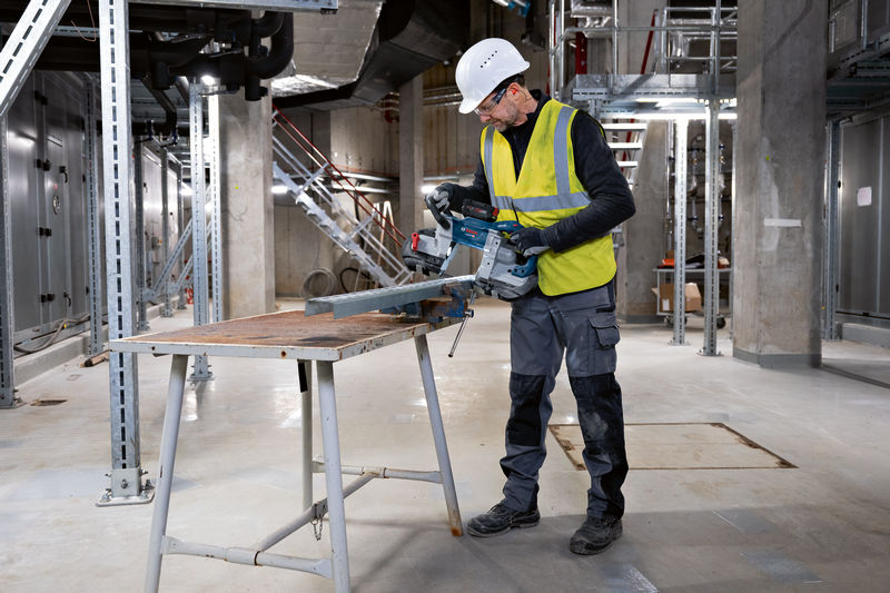 Eine Person mit Schutzausrüstung schneidet einen Metallträger mit einer Akku-Bandsäge. Anwendungsbild