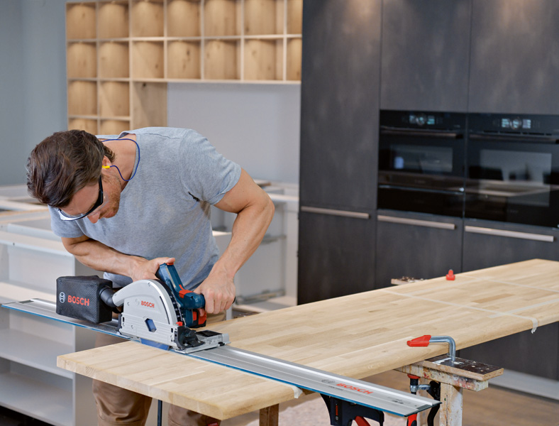 Eine Person mit Sicherheitsausrüstung schneidet in einer modernen Küche mit einer Tauchsäge ein Holzbrett. Anwendungsbild