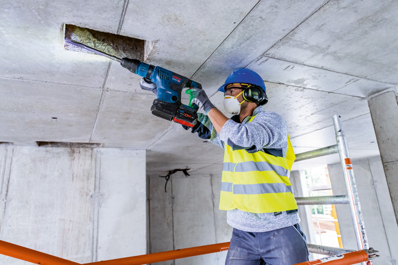 Eine Person mit Schutzausrüstung bohrt mit einem Akku-Bohrhammer in eine Betondecke. Anwendungsbild