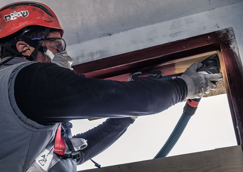 Eine Person mit Schutzausrüstung schleift mit einem Elektrowerkzeug einen hölzernen Fensterrahmen. Anwendungsbild