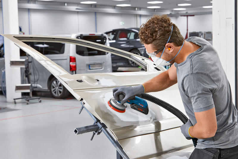 Eine Person mit Schutzausrüstung schleift in einer Autowerkstatt eine Autotür mit einem elektrischen Schleifgerät. Anwendungsbild