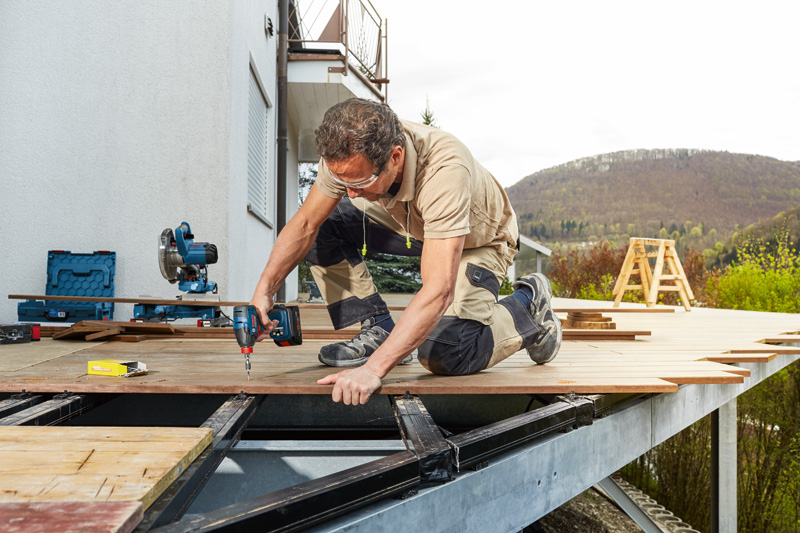 Eine Person mit Sicherheitsausrüstung bohrt Terrassendielen in einen Terrassenrahmen im Freien. Anwendungsbild