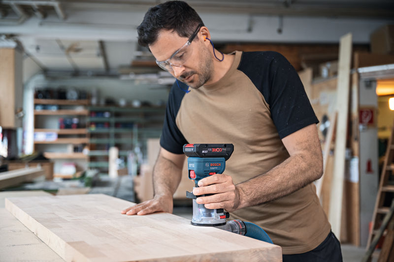 Eine Person mit Schutzausrüstung bedient eine kabellose Handfräse auf einem Holzbrett. Anwendungsbild