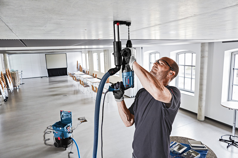 Eine Person mit Schutzausrüstung bohrt mit einer Staubabsauganlage in eine Decke. Anwendungsbild