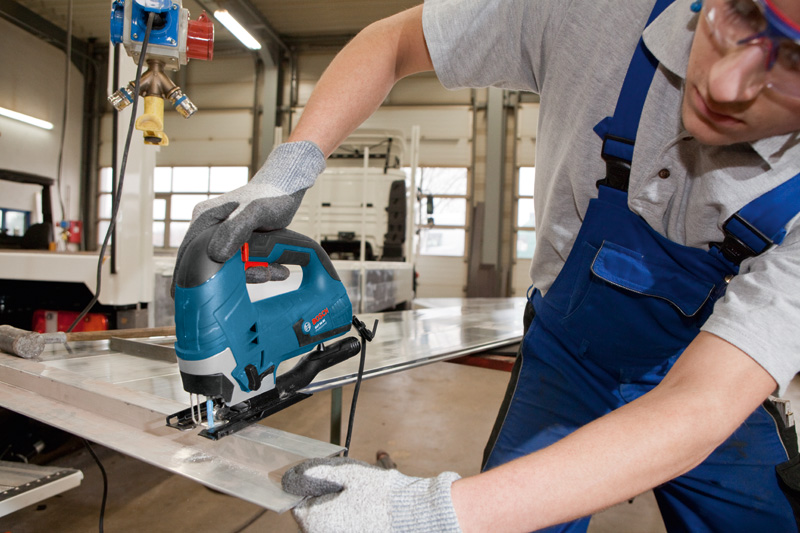 Eine Person mit Sicherheitsausrüstung führt eine Stichsäge, um ein Metallblech zu schneiden. Anwendungsbild