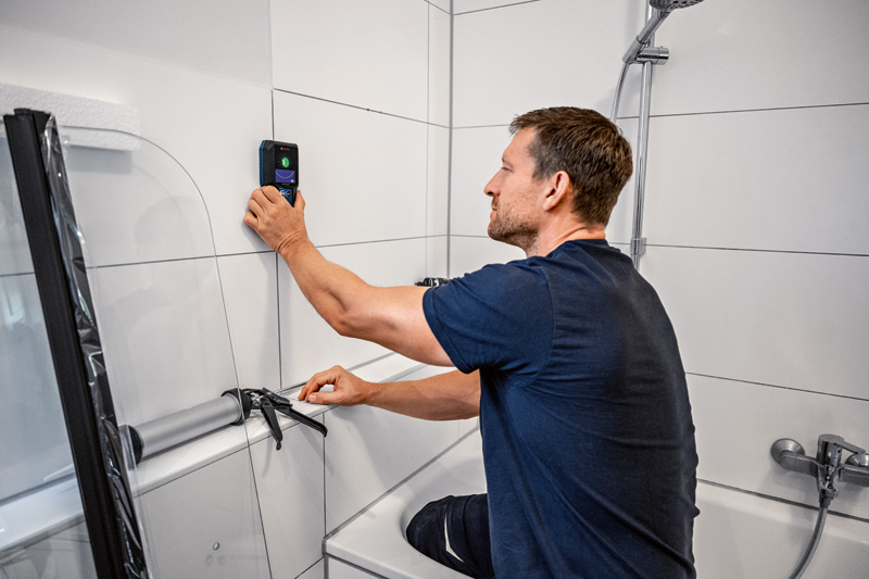 Eine Person verwendet einen Wanddetektor auf weißen Badezimmerfliesen in der Nähe einer Badewanne. Anwendungsbild