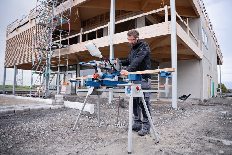 Eine Person mit Sicherheitsausrüstung schneidet auf einer Baustelle Holz mit einer Akku-Gehrungssäge. Anwendungsbild