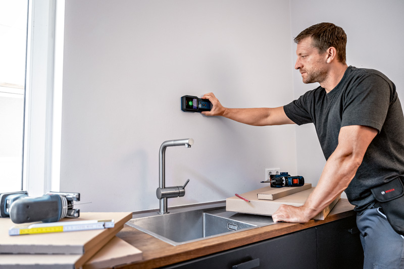 Eine Person scannt mit einem digitalen Detektor eine Küchenwand über einer Spüle. Anwendungsbild
