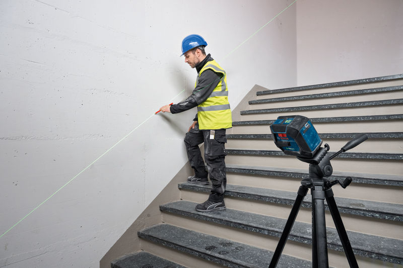 Eine Person mit Sicherheitsausrüstung markiert mit einem Lasernivelliergerät eine Wand auf einer Treppe. Anwendungsbild