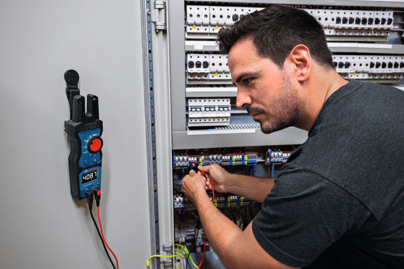 Eine Person überprüft elektrische Leitungen in einem Bedienfeld mit einem digitalen Multimeter. Anwendungsbild