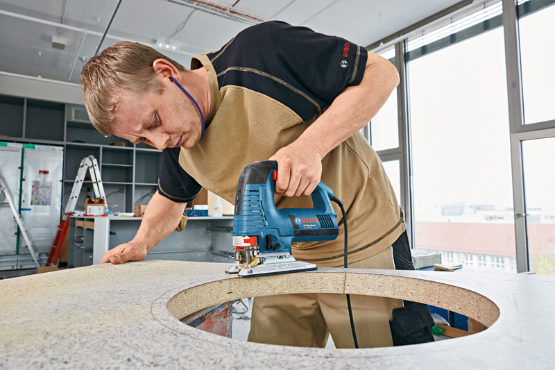 Eine Person mit Schutzausrüstung schneidet mit einer Stichsäge eine kreisförmige Öffnung in eine Arbeitsplatte. Anwendungsbild