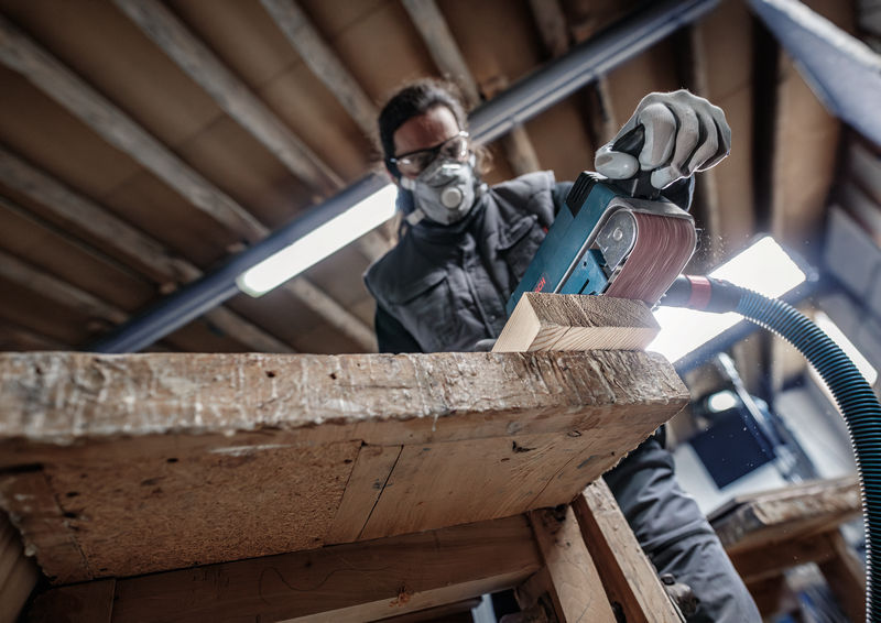 Eine Person mit Sicherheitsausrüstung schleift einen Holzbalken mit einem elektrischen Bandschleifer. Anwendungsbild