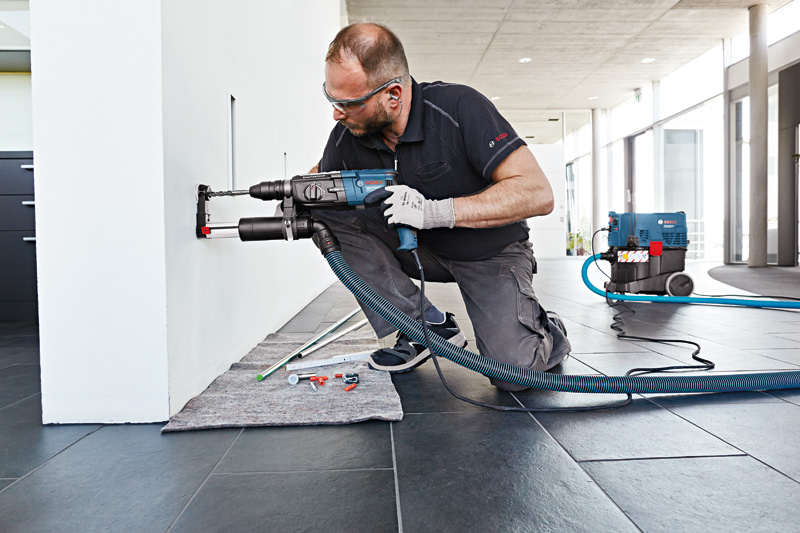 Eine Person mit Schutzausrüstung bohrt mit einem Staubabsaugaufsatz in eine Wand. Anwendungsbild