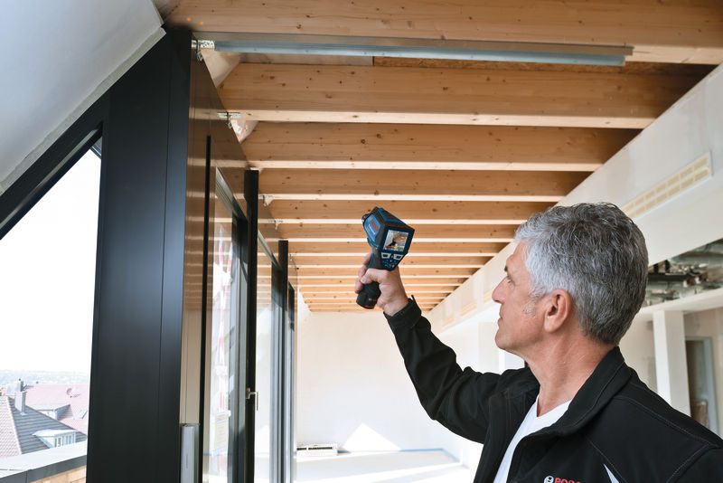 Eine Person tastet in einem hellen Raum mit einem Thermodetektor eine Holzdecke ab. Anwendungsbild
