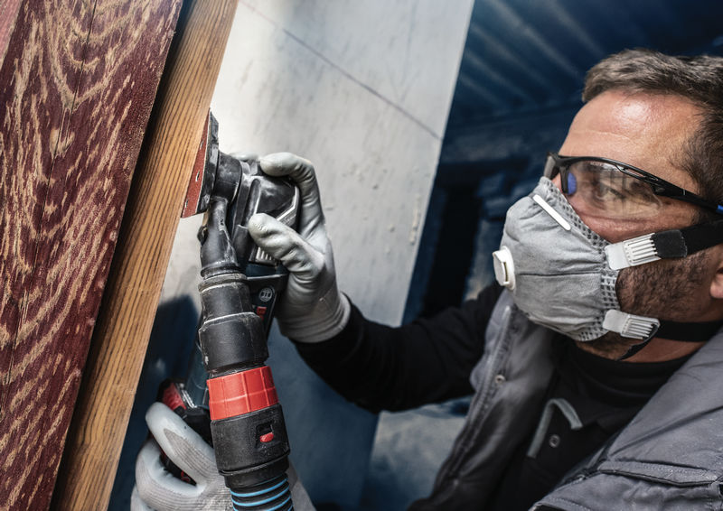 Ein Arbeiter in Sicherheitsausrüstung schleift ein Holzbrett mit einem Elektrowerkzeug. Anwendungsbild