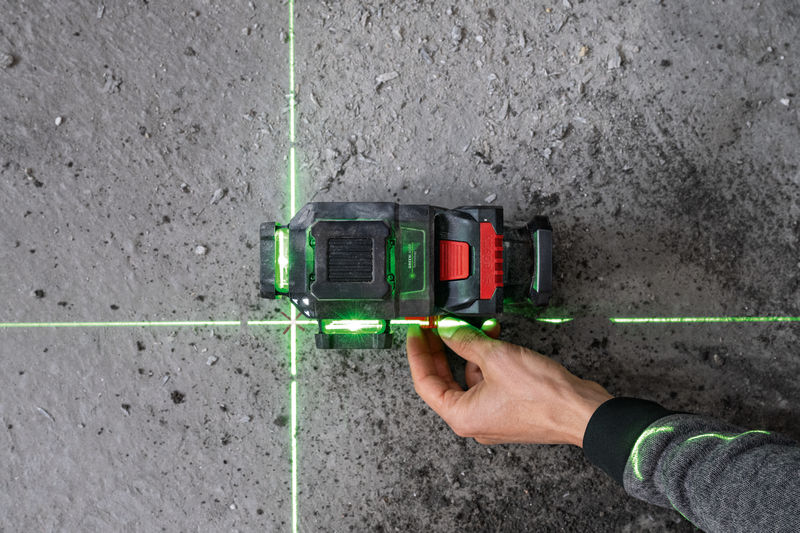 Eine Person richtet ein Lasernivelliergerät aus, das grüne Linien auf einen Betonboden projiziert. Anwendungsbild