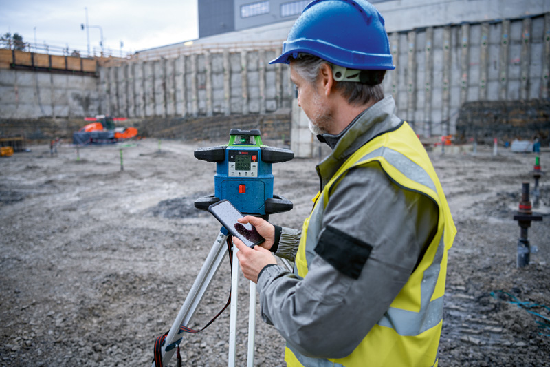 Eine Person mit Sicherheitsausrüstung bedient auf einer Baustelle ein Lasernivelliergerät. Anwendungsbild
