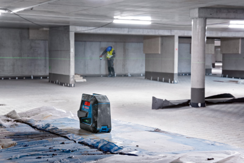 Lasernivelliergerät projiziert grüne Linien in eine Garage, im Hintergrund eine Person mit Sicherheitsausrüstung. Anwendungsbild