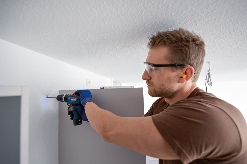 Eine Person mit Schutzausrüstung bohrt beim Montieren von Möbeln in eine Wand. Anwendungsbild