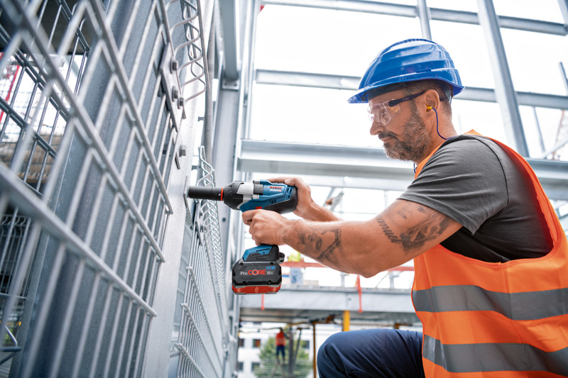 Eine Person mit Sicherheitsausrüstung befestigt mit einem Akku-Schlagschrauber einen Metallzaun. Anwendungsbild