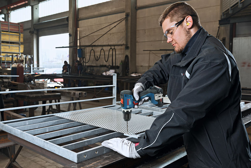 Eine Person mit Sicherheitsausrüstung bohrt in einer Werkstatt Metallgitter auf einer Werkbank. Anwendungsbild