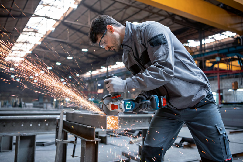 Eine Person mit Schutzausrüstung schleift in einer Fabrik Metall mit einem Akku-Winkelschleifer. Anwendungsbild