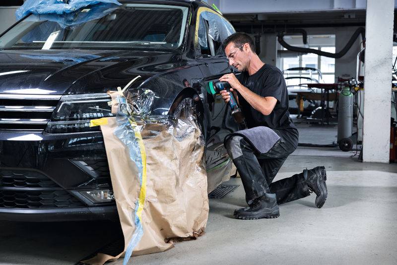 Eine Person mit Sicherheitsausrüstung verwendet in einer Autowerkstatt eine Poliermaschine an einem Kotflügel. Anwendungsbild