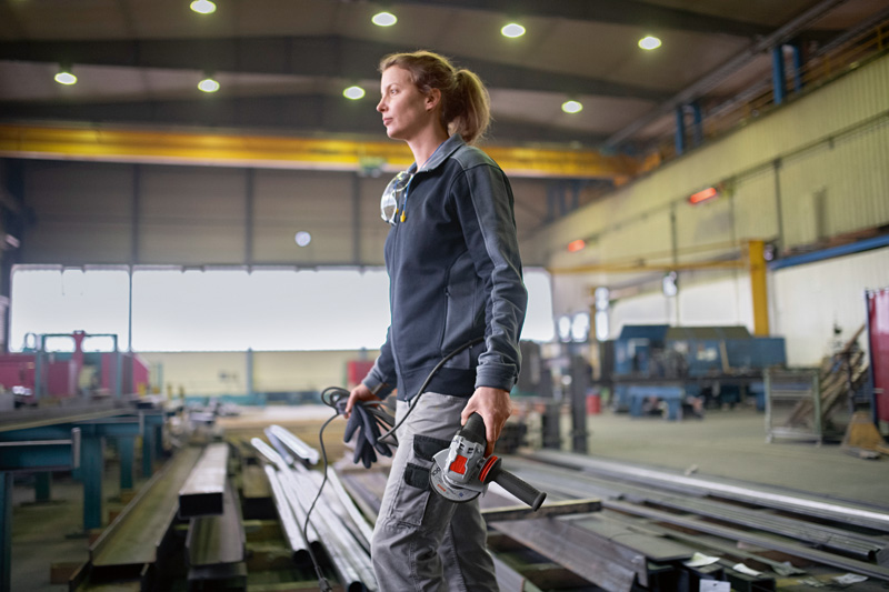 Eine Person mit Schutzausrüstung trägt einen Winkelschleifer in einer großen Industriewerkstatt. Anwendungsbild