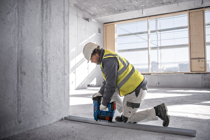 Eine Person mit Sicherheitsausrüstung befestigt mit einem Akku-Betonnagler eine Metallschiene am Boden. Anwendungsbild
