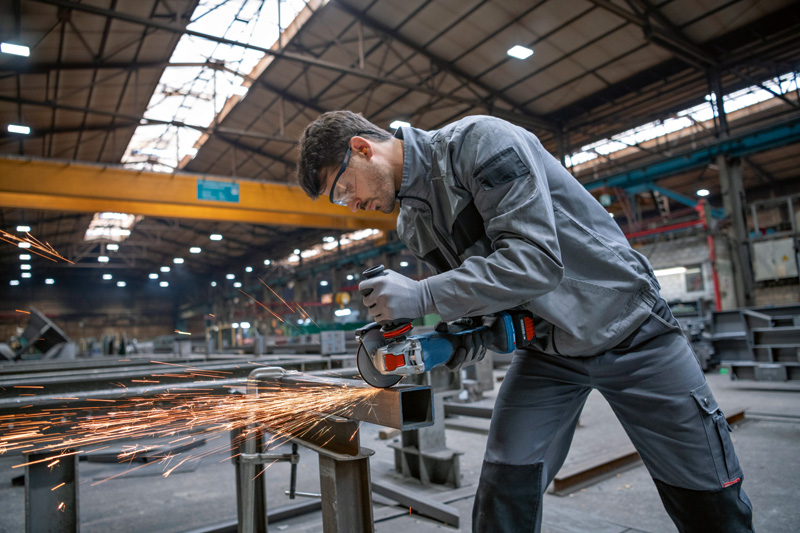 Eine Person mit Schutzausrüstung schleift in einer großen Werkstatt Metall und erzeugt dabei Funken. Anwendungsbild