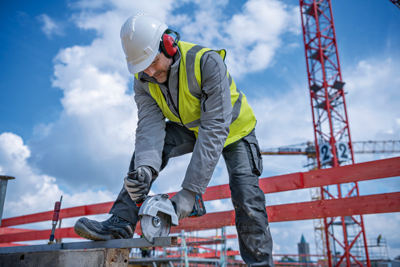Eine Person mit Sicherheitsausrüstung verwendet einen Akku-Winkelschleifer, um auf einer Baustelle Metall zu schneiden. Anwendungsbild