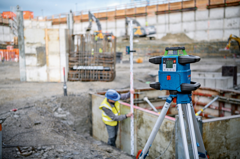 Lasernivelliergerät auf einem Stativ auf einer Baustelle, Person trägt Sicherheitsausrüstung. Anwendungsbild