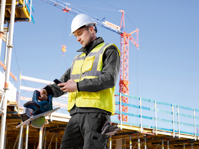 Eine Person mit Sicherheitsausrüstung überprüft auf einer Baustelle ein Telefon, während sie eine Kreissäge hält. Anwendungsbild
