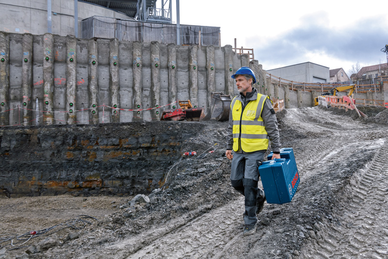 Eine Person mit Sicherheitsausrüstung trägt einen blauen Werkzeugkoffer auf einer schlammigen Baustelle. Anwendungsbild