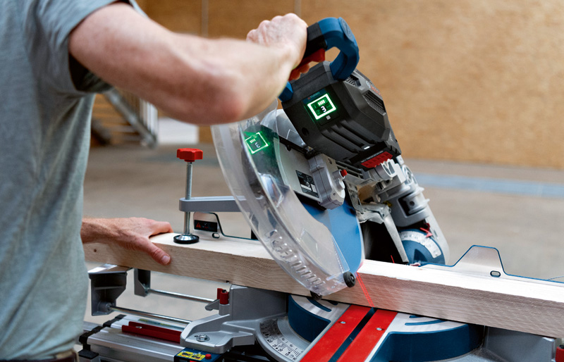 Eine Person bedient eine kabellose Gehrungssäge, um ein Holzbrett auf einem Werkstatttisch zu schneiden. Anwendungsbild