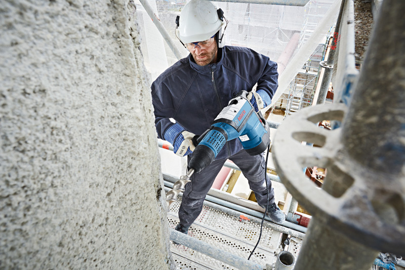 Eine Person mit Schutzausrüstung bohrt auf einem Gerüst mit einem Bohrhammer in Beton. Anwendungsbild