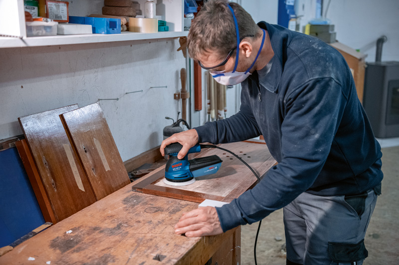 Eine Person mit Schutzausrüstung schleift ein Holzbrett mit einem Exzenterschleifer. Anwendungsbild