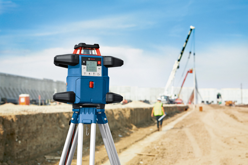 Auf einem Stativ auf einer Baustelle aufgestelltes Lasernivelliergerät, in der Nähe eine Person mit Sicherheitsausrüstung. Anwendungsbild