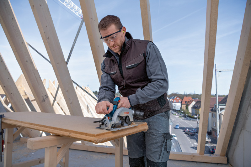 Eine Person mit Sicherheitsausrüstung schneidet auf einer Baustelle mit einer Kreissäge ein Holzbrett. Anwendungsbild