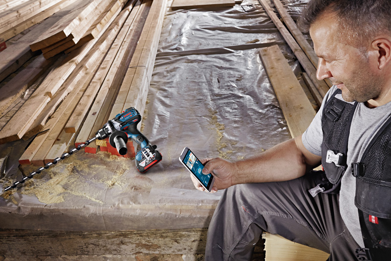 Eine Person mit Sicherheitsausrüstung steuert auf einer Baustelle eine Akku-Bohrmaschine mit einem Smartphone. Anwendungsbild