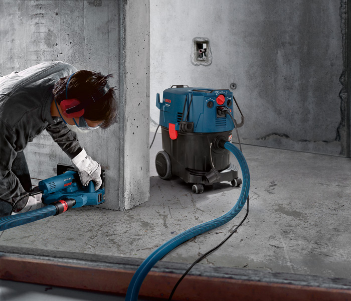 Eine Person mit Schutzausrüstung schleift Beton mit einem über einen Schlauch angeschlossenen Staubabsauger. Anwendungsbild