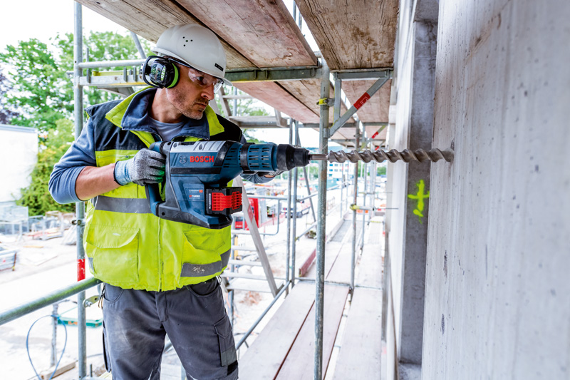 Eine Person mit Sicherheitsausrüstung bohrt auf einer Baustelle in eine Betonwand. Anwendungsbild