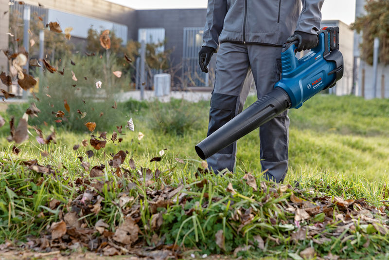 Eine Person mit Sicherheitsausrüstung entfernt mit einem Akku-Laubbläser Blätter vom Gras. Anwendungsbild